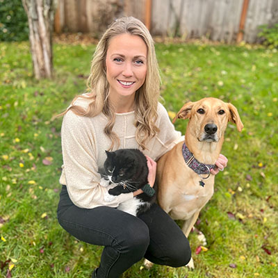 A woman is sitting on the grass, smiling at the camera. She is holding a black and white cat and sitting beside a tan dog wearing a colorful collar. A wooden fence and trees are in the background.