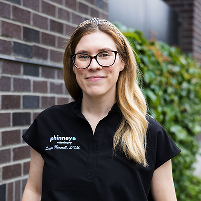 A person with long blonde hair, wearing glasses, a black shirt with a veterinary logo, and a leopard print headband, stands in front of a brick wall and greenery.