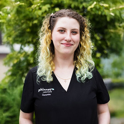 A person with curly, light hair stands outdoors, wearing a black shirt with "Phinney Veterinary" and "Samantha" written on it. The background features green, leafy trees.